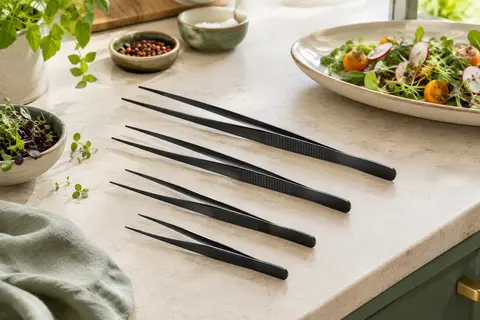 Quatre pincettes de cuisine noires en acier inoxydable, deux longueurs et pointes fines sur un plan clair.